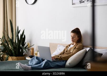 A young woman enjoys a chill day in bed with her laptop, surrounded by a peaceful apartment vibe. Stock Photo