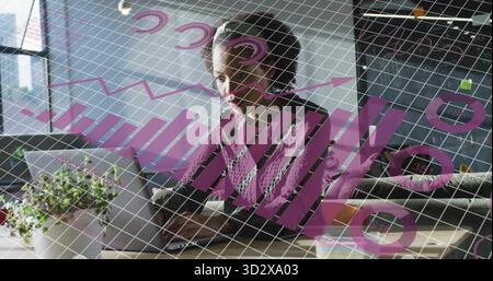 Analyzing mid-adult woman reviewing charts on laptop at desk in office, with plant, sticky notes Stock Photo