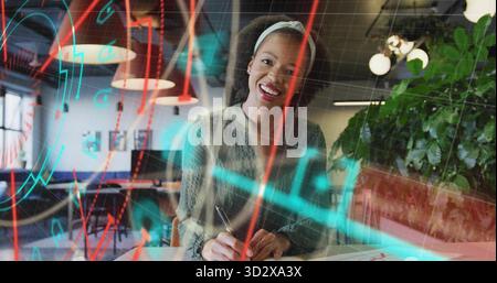 Writing office worker wearing sweater at open-plan desk, using pen on notebook with overlaid charts Stock Photo