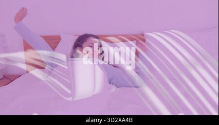 Stretching Indian woman wearing blue pajamas in bedroom with bed duvet pillows nightstand lamp Stock Photo