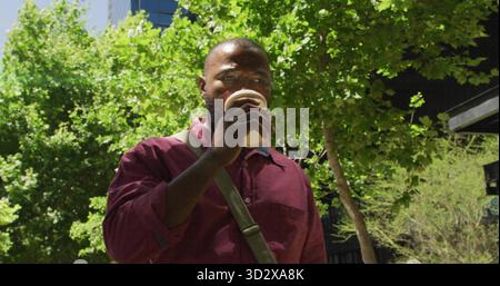 Drinking man holding paper coffee cup sporting maroon shirt and shoulder bag on tree-lined plaza Stock Photo