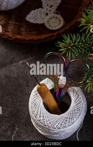 Close-up of colorful embroidery scissors and a crochet hook in a spool of white cotton thread on a dark stone background. Handicraft, crochet, and DIY Stock Photo