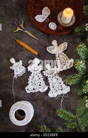 Flat lay composition with handmade white crochet Christmas angels in various stages of completion, crochet hook, scissors, and burning candle on dark Stock Photo
