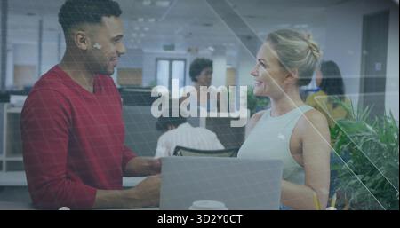 Standing colleagues wearing red and green tops discussing at desk in open-plan office, with laptop Stock Photo
