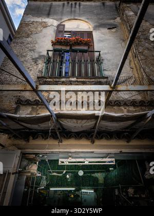 An old building on a Belgrade street with cracked walls and a small balcony featuring closed wooden shutters and red flowers, viewed from below throug Stock Photo