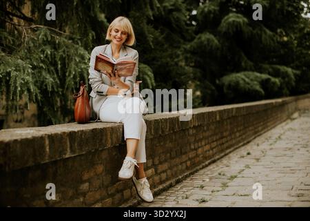 A blonde woman wearing a blazer and white pants sits on a stone wall outside reading. She is smiling and has a brown purse next to her in a park setti Stock Photo