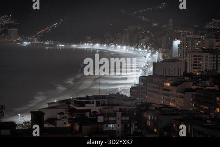 Telephoto long exposure of Ipanema beach at night, illuminated shoreline leading toward Arpoador and distant Leblon, blurred ocean wave motion and cit Stock Photo