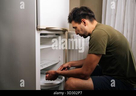 Side view of man sitting on stool, defrosting refrigerator and cleaning interior, performing essential maintenance in kitchen Stock Photo