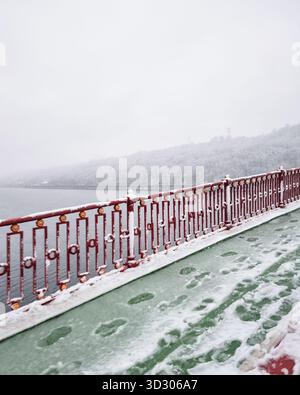Empty pedestrian bridge over the river covered with slush snow. Red barrier of pedestrian footbridge. Snow-capped trees on the hill in the background. Stock Photo