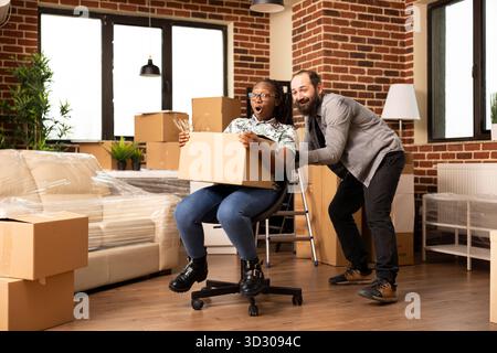 Young multiethnic couple enjoying playful moments in new home, laughing and riding on chair surrounded by cardboard boxes. Happy boyfriend and girlfriend celebrate moving day with fun and excitement. Stock Photo