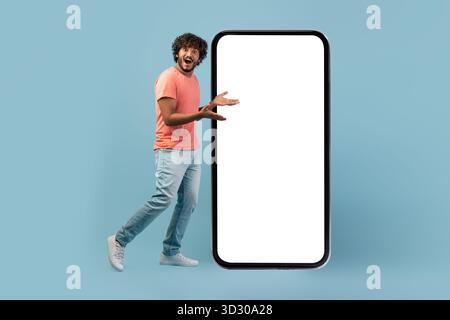 Curly Bearded Guy Amazed by Large Smartphone With Blank Screen in Blue Studio Setup Stock Photo