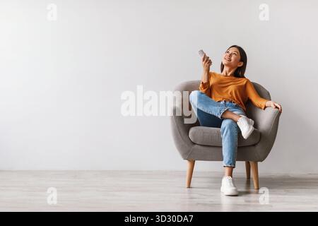 Young Woman Relaxes With Remote in Armchair Under Air Conditioner Stock Photo