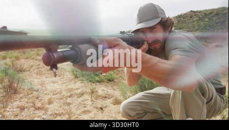 Kneeling male aiming rifle with scope on arid desert terrain, with ammunition belt and baseball cap Stock Photo