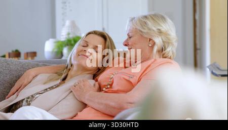 Relaxing adult daughter leaning on mother's shoulder in living room, with gray couch, vases, plants Stock Photo