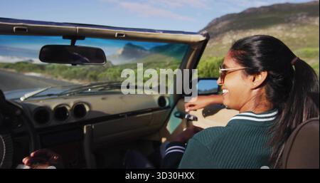 Riding convertible car on coastal road with passenger holding handle wearing sunglasses, copy space Stock Photo