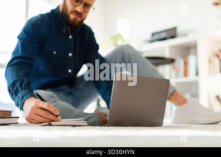 Young Man Studying at Home With Laptop and Notebook in a Cozy, Bright Living Room Space During the Day Stock Photo