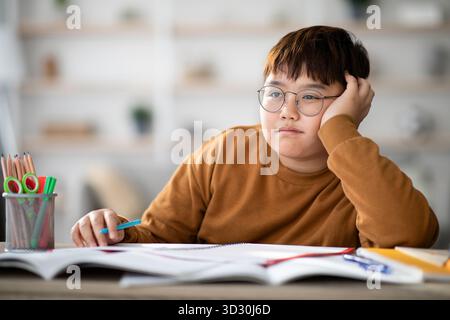 Chubby Asian School Boy Daydreaming While Doing Homework at a Desk in His Home Stock Photo