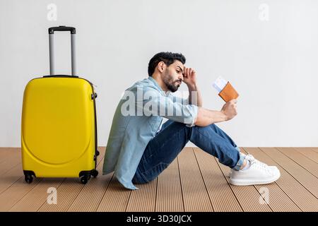 Man Sitting on the Floor Beside Yellow Suitcase, Looking Anxious While Holding Travel Documents Before Departure Stock Photo