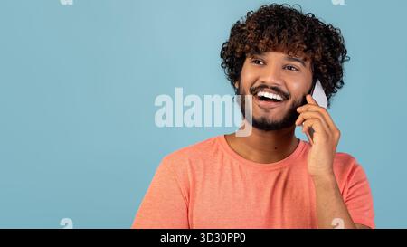 Positive Indian Guy Enjoying a Phone Conversation on a Blue Background While Smiling Stock Photo