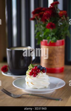 A delicate meringue dessert topped with fresh berries sits on a plate next to a black coffee cup. A pot of red flowers adds color to the warm scene. Stock Photo