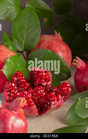 Pomegranates are displayed on a textured surface, showing ripe fruits and several seeds spilling from a half-opened fruit. Fresh green leaves add cont Stock Photo