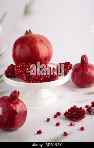 Red pomegranates are displayed on a white pedestal. Some are whole while others are cut open, revealing shiny red arils scattered nearby. The setting Stock Photo