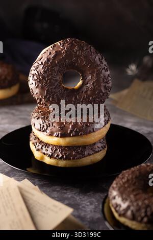 Three thick chocolate-covered donuts are stacked neatly on a black plate. Surrounding them are vintage papers, adding a warm, nostalgic touch to the s Stock Photo