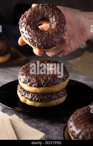 A hand lifts a chocolate-covered donut above a stack of similar treats on a dark plate. The scene is set in a cozy, inviting kitchen. Stock Photo