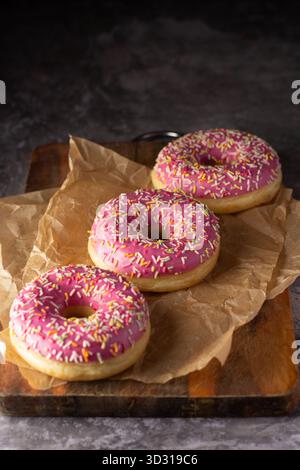 Three pink frosted donuts topped with colorful sprinkles rest on parchment paper on a wooden board in a warm dessert shop. The atmosphere is inviting Stock Photo