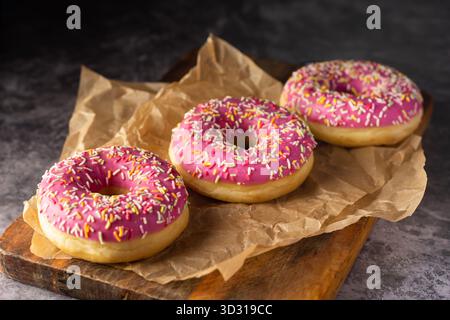 Three delicious donuts topped with bright pink icing and colorful sprinkles sit on crumpled parchment paper on a wooden board. The warm, inviting kitc Stock Photo