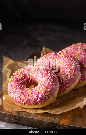 Three bright pink donuts with colorful sprinkles are arranged neatly on brown parchment paper. The soft lighting highlights their texture, making them Stock Photo