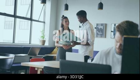 Discussing project coworkers reviewing green folder at laptop in open plan office, with lamps Stock Photo