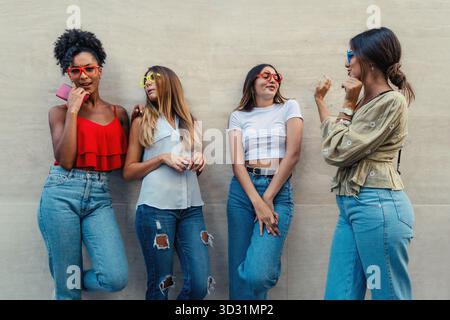 A happy group of four young female friends wear colorful glasses and laugh together against an urban wall. Stock Photo