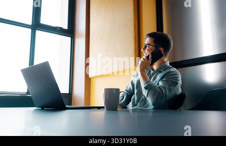 A young adult man wearing glasses, using a smartphone, and seated at a desk with a laptop in an office setting, appearing focused and engaged. Stock Photo