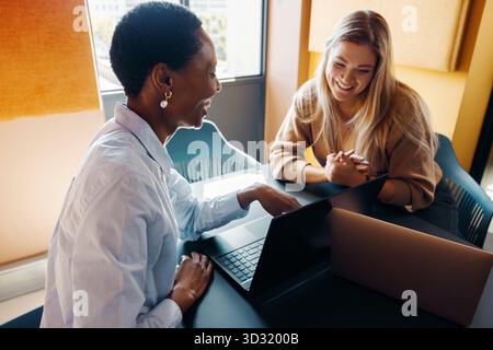 Two young women smile while working together at a table, using laptops in an office. This depicts a professional and cooperative atmosphere. Stock Photo