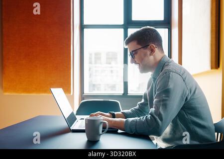 A young adult man wearing glasses focuses on his laptop in a well-lit workplace, accompanied by a cup of coffee, appearing diligent and engaged. Stock Photo