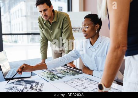 A multicultural team of designers meeting and discussing building plans in a bright office. One woman shows architectural designs to two colleagues. Stock Photo