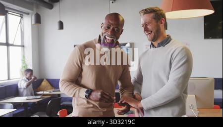 Laughing coworkers exchanging ring in shared workspace, wearing pullovers with laptop smartwatch Stock Photo