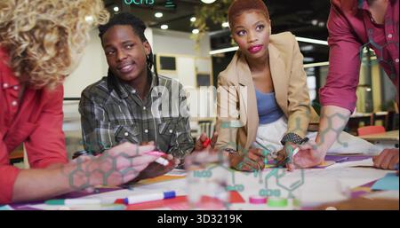 Collaborating professionals leaning over diagrams on office table with markers and chemical overlay Stock Photo