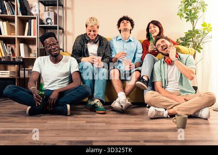 A happy, multicultural group of young friends (including a Black man) laugh while watching a movie, eating popcorn, and drinking beer in a cozy living Stock Photo