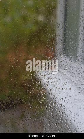 Rain on the window and view out of it: rainy day, white outer window sill and raindrops on the glass, blurred colorful background of a garden Stock Photo
