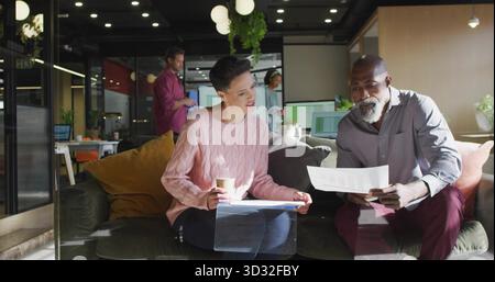 Reviewing paperwork mature colleagues sitting on dark sofa in modern office lounge, with coffee cup Stock Photo