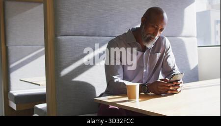 Holding smartphone, senior Black man wearing shirt sitting at modern lounge table with coffee cup Stock Photo