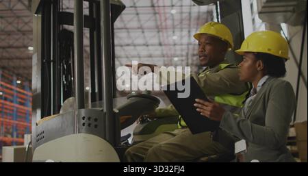 Pointing forklift operator directing supervisor inside warehouse, with clipboard and metal racks Stock Photo