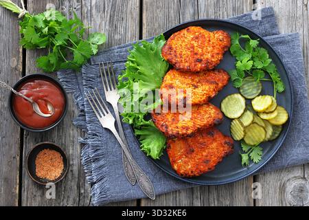 Paprika Seasoned Pork Chops on plate with lettuce and sliced dill pickles on rustic wood table with forks, tomato sauce and fresh cilantro, horizontal Stock Photo