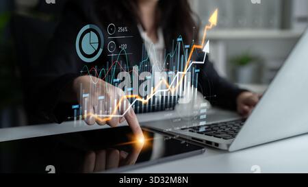 A businesswoman is analyzing financial data with digital graphs on her laptop and interacting with a tablet in a modern office, showcasing strategic i Stock Photo