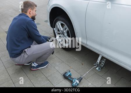 Man changing car tire using electric drill and jack on city sidewalk. DIY auto repair, maintenance, seasonal tire change, mechanical work, vehicle Stock Photo