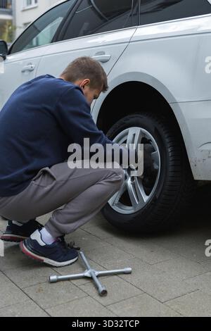Man changing car tire using electric drill and jack on city sidewalk. DIY auto repair, maintenance, seasonal tire change, mechanical work, vehicle Stock Photo
