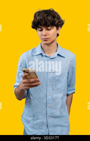 A young man with curly hair stands, looking down and texting or scrolling on his smartphone, isolated on a vibrant yellow background. Stock Photo