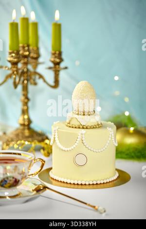 Small easter cake decorated with white chocolate egg, golden beads and white pearls, lit green candles and teacup in background, festive atmosphere Stock Photo